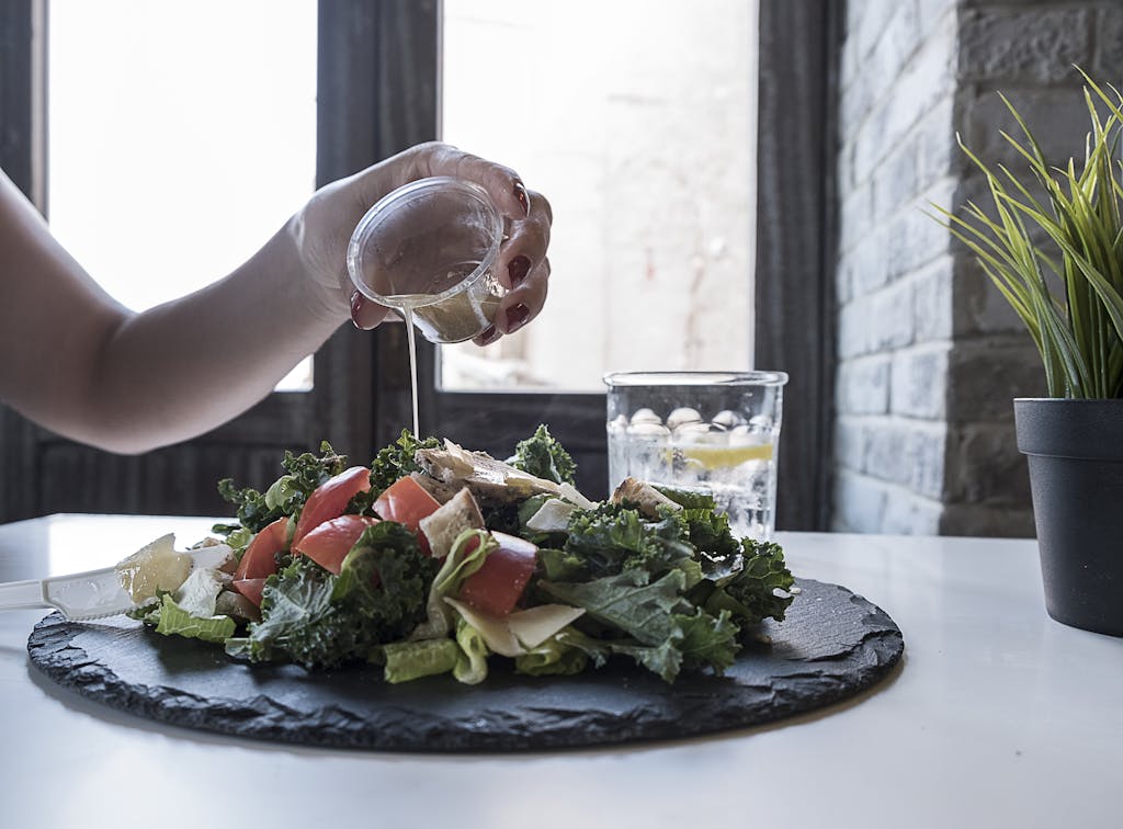 Hand pouring dressing over fresh salad with kale and tomatoes in a modern Dubai restaurant.