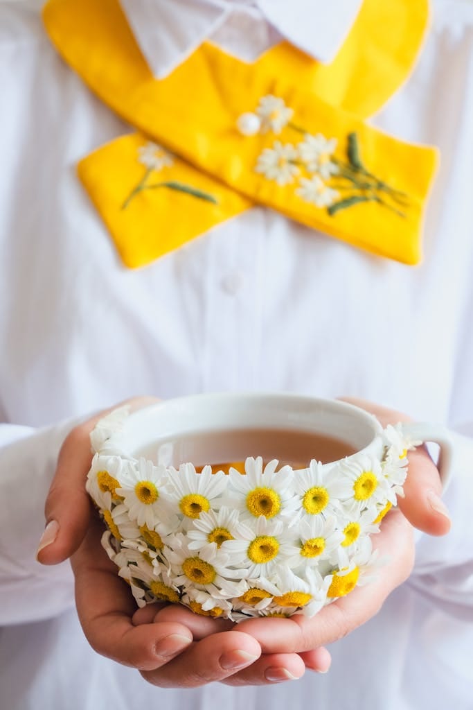A woman holds a floral tea cup decorated with daisies, wearing a white shirt with yellow daisy embroidery.